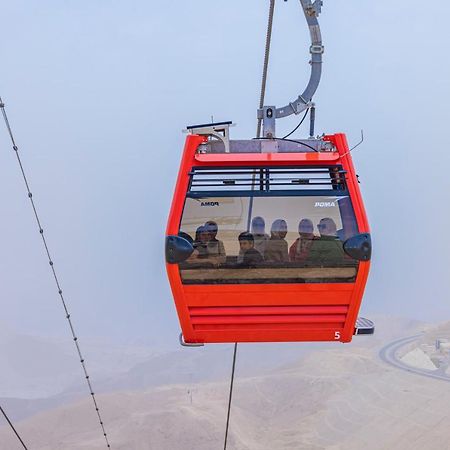 Tolip Resort El Galala Majestic Ajn-Szuhna Kültér fotó The photo shows a red gondola lift suspended in the air, traveling along cables. Inside the gondola, a group of people can be seen. The background features a hazy landscape, possibly mountains or hills, indicating an elevated area. The scene conveys 