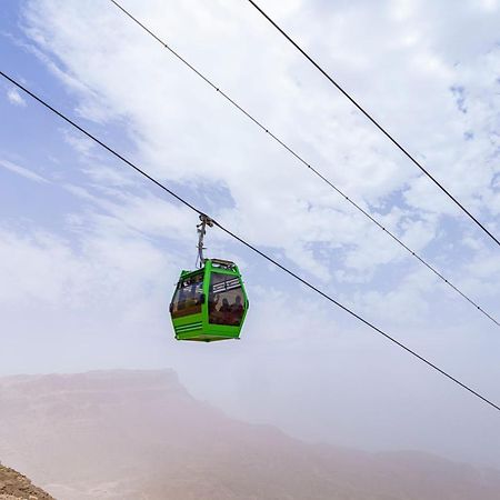 Tolip Resort El Galala Majestic Ajn-Szuhna Kültér fotó The photo shows a green cable car suspended in the air, traveling along overhead cables. The backdrop features a hazy landscape, likely mountainous or elevated terrain, with a cloudy sky. The scene suggests a scenic transportation method, possibly us