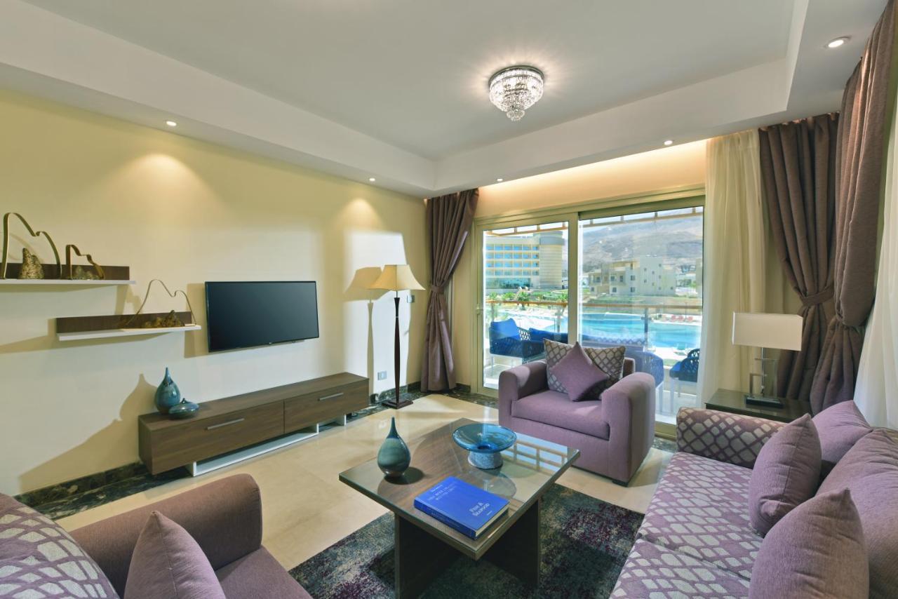 Tolip Resort El Galala Majestic Ajn-Szuhna Kültér fotó The photo shows a modern living room that features a stylish and cozy design. There are two purple sofas with decorative cushions arranged around a coffee table. The coffee table has a blue book and a decorative item on it. A flat-screen TV is mounte