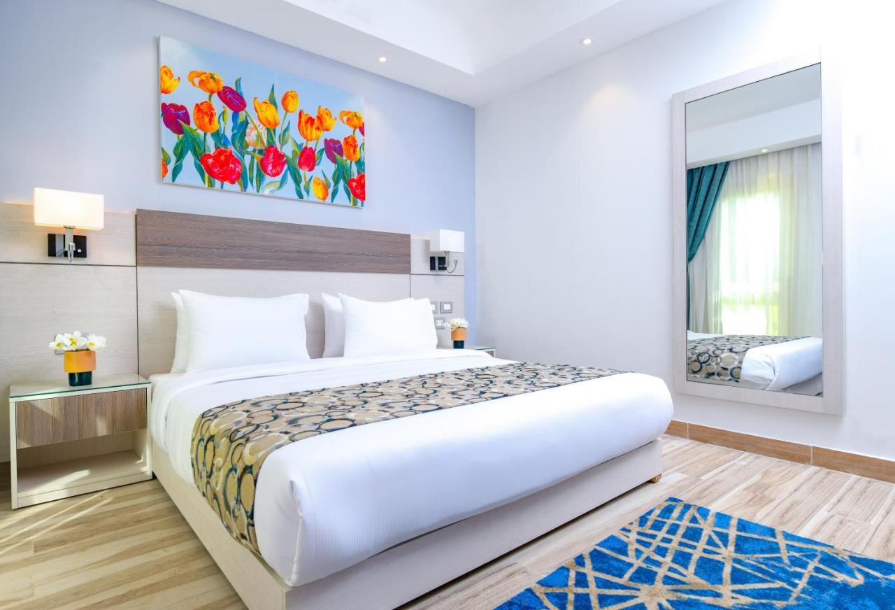 Tolip Resort El Galala Majestic Ajn-Szuhna Kültér fotó The photo shows a modern hotel bedroom. It features a large bed with white linens and a decorative throw blanket. The room has light wooden flooring and is bright, with ample natural light coming from a window. There is a colorful painting of flowers