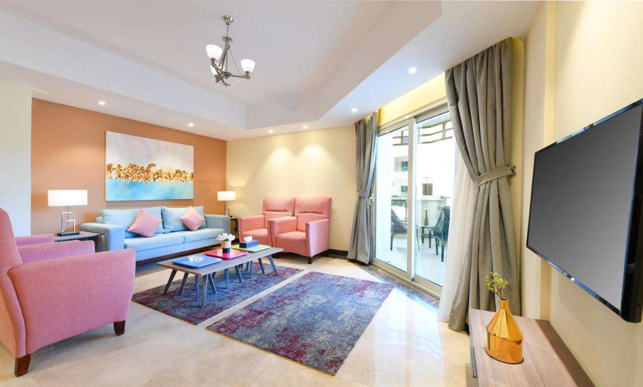 Tolip Resort El Galala Majestic Ajn-Szuhna Szoba fotó The photo shows a modern living room that features a cozy and stylish design. There is a light blue sofa adorned with pastel pink cushions, accompanied by two pink armchairs. A coffee table sits in front, placed on a colorful area rug that adds warmt