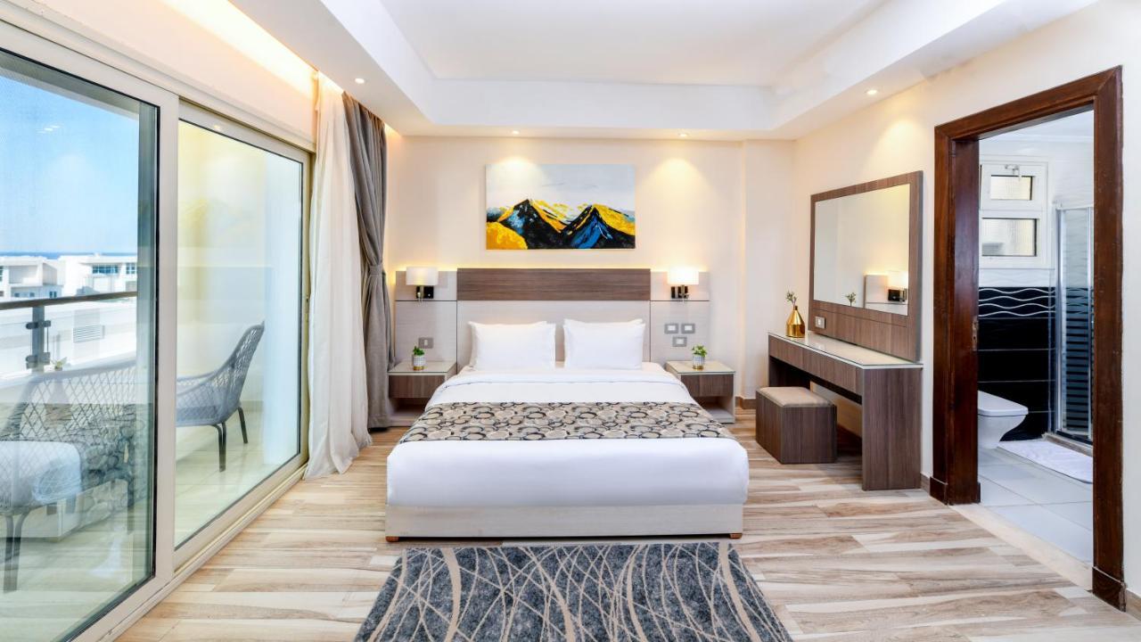 Tolip Resort El Galala Majestic Ajn-Szuhna Kültér fotó The photo shows a modern hotel room featuring a large bed with white linens and a decorative throw. The room has a neutral color palette, accented with warm lighting. There is a piece of artwork above the bed depicting mountains. To the left, there a