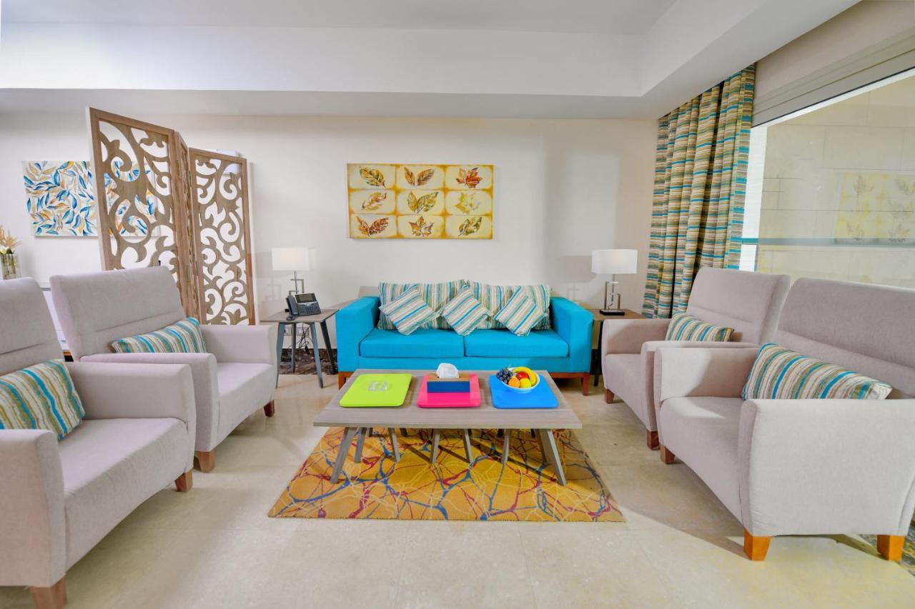 Tolip Resort El Galala Majestic Ajn-Szuhna Kültér fotó The photo shows a stylish and colorful living room. It features a bright blue sofa with patterned cushions, complemented by two light gray armchairs. In front of the sofa, there's a wooden coffee table with vibrant trays in pink, green, and yellow. A
