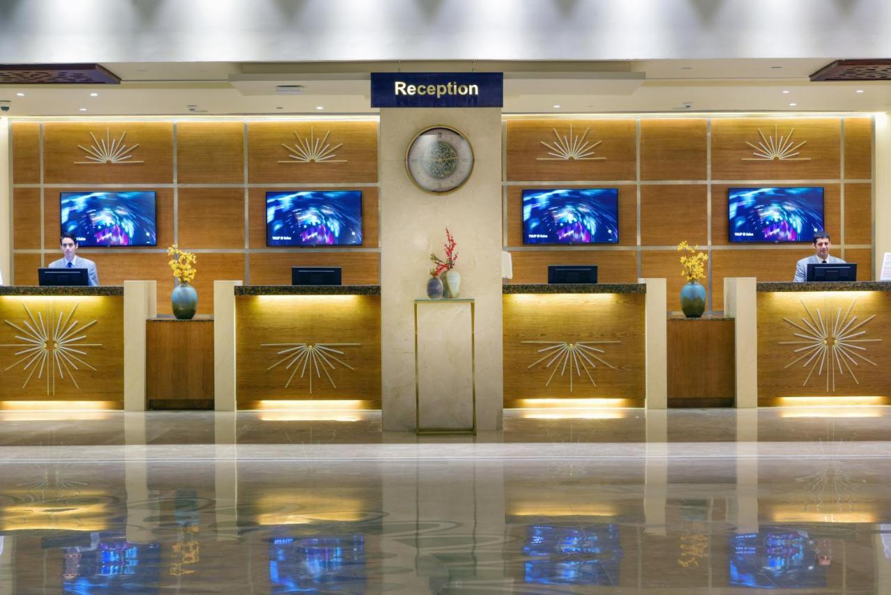 Tolip Resort El Galala Majestic Ajn-Szuhna Kültér fotó The image shows a hotel reception area. There are several reception desks lined up against a wall with decorative paneling. Each desk has a staff member, and above the desks, there are screens displaying information. A clock is visible on the wall, a
