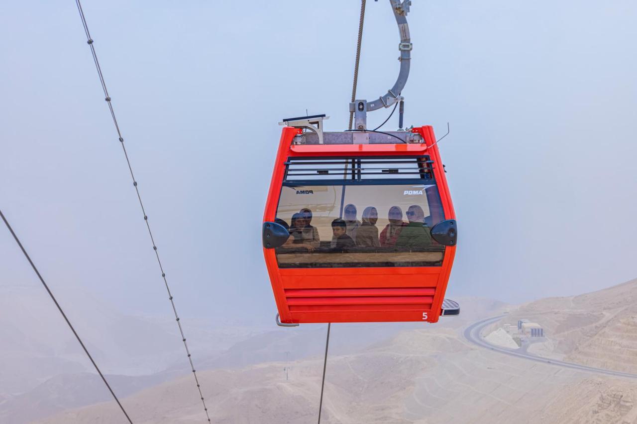 Tolip Resort El Galala Majestic Ajn-Szuhna Kültér fotó The photo shows a red gondola lift suspended in the air, traveling along cables. Inside the gondola, a group of people can be seen. The background features a hazy landscape, possibly mountains or hills, indicating an elevated area. The scene conveys 