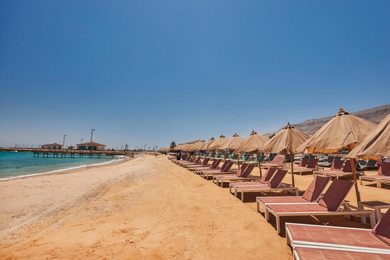 Tolip Resort El Galala Majestic Ajn-Szuhna Kültér fotó The photo shows a scenic beach with a stretch of golden sand under a clear blue sky. There are several beach loungers arranged in rows, each accompanied by thatched umbrellas providing shade. In the background, there is a wooden pier extending into t