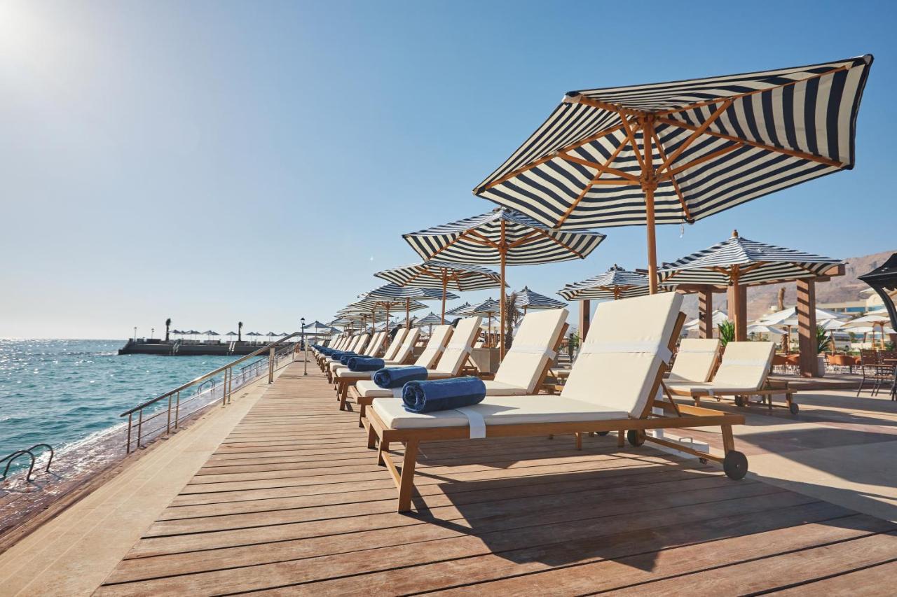 Tolip Resort El Galala Majestic Ajn-Szuhna Kültér fotó The photo depicts a serene beachside setting featuring a row of sun loungers arranged on a wooden deck. Each lounger is equipped with a folded towel and shaded by striped umbrellas. In the background, the water sparkles under bright sunlight, and the