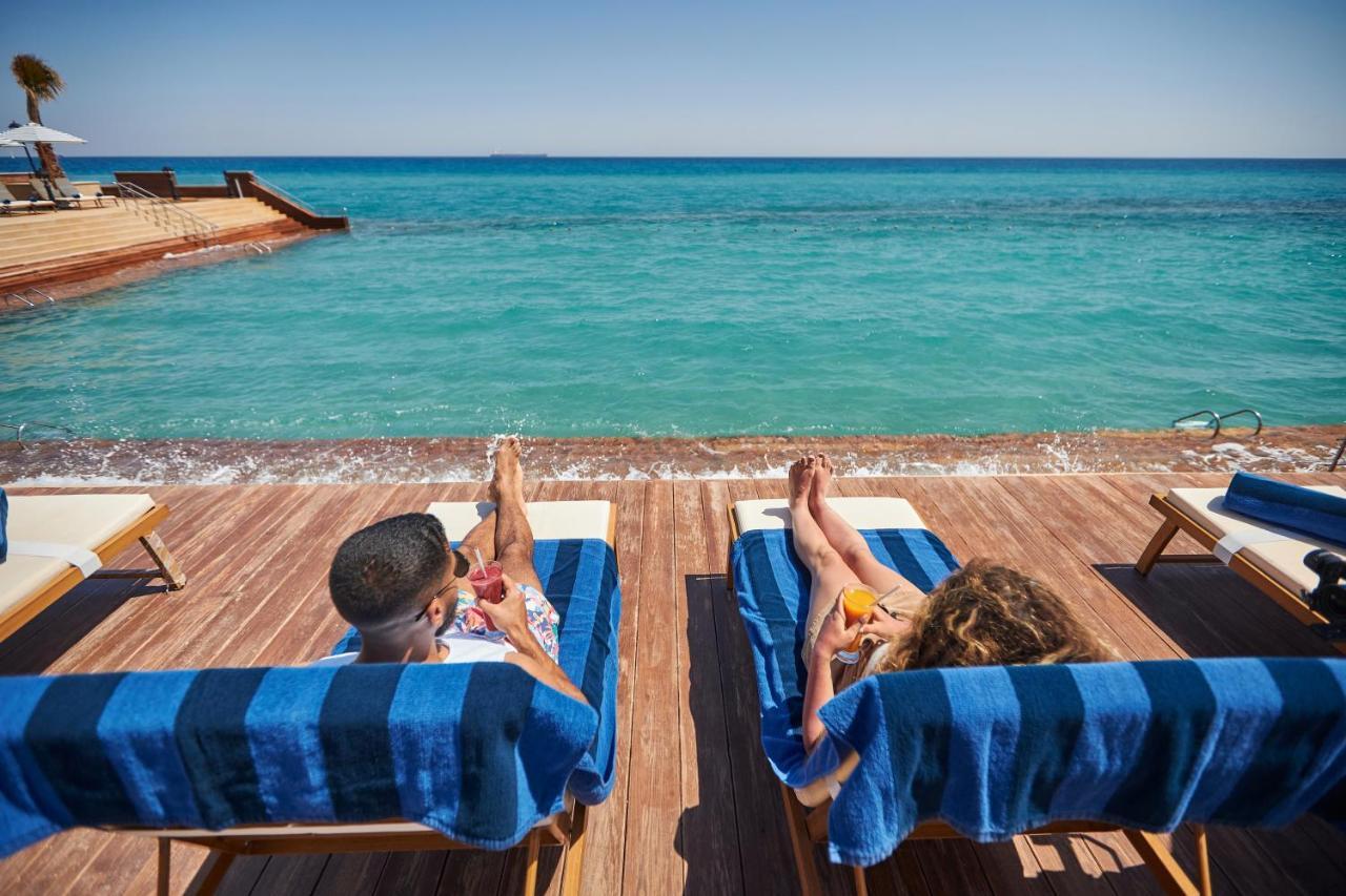 Tolip Resort El Galala Majestic Ajn-Szuhna Kültér fotó The photo depicts two people relaxing on lounge chairs by the waterfront. They are positioned on a wooden deck with a view of the clear, turquoise water. Both individuals are reclining, enjoying the sun, and each has a drink in hand. The scene convey