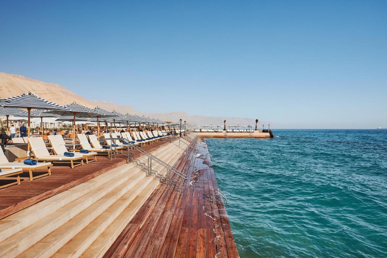 Tolip Resort El Galala Majestic Ajn-Szuhna Kültér fotó The photo shows a seaside area featuring a wooden deck that extends into the water. On the left side, there are several lounge chairs arranged neatly under shade structures, suggesting a relaxing atmosphere. The ocean is visible in the foreground, wi