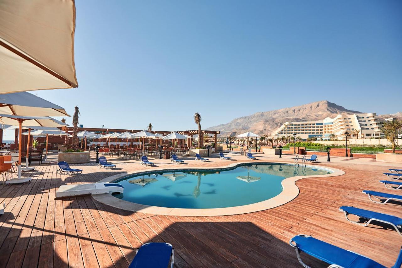 Tolip Resort El Galala Majestic Ajn-Szuhna Kültér fotó The photo shows a swimming pool area surrounded by sun loungers and umbrellas. The pool has a unique, curvy shape and is situated on a wooden deck. In the background, there are several buildings, likely part of a resort or hotel, and a mountainous la