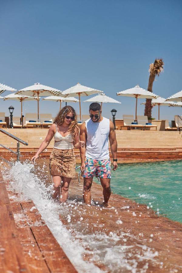 Tolip Resort El Galala Majestic Ajn-Szuhna Kültér fotó The photo depicts a couple walking along a poolside area. They are enjoying a sunny day near a swimming pool, with splashes of water around them. The woman is wearing a light-colored top and a patterned skirt, while the man is dressed in a sleeveless