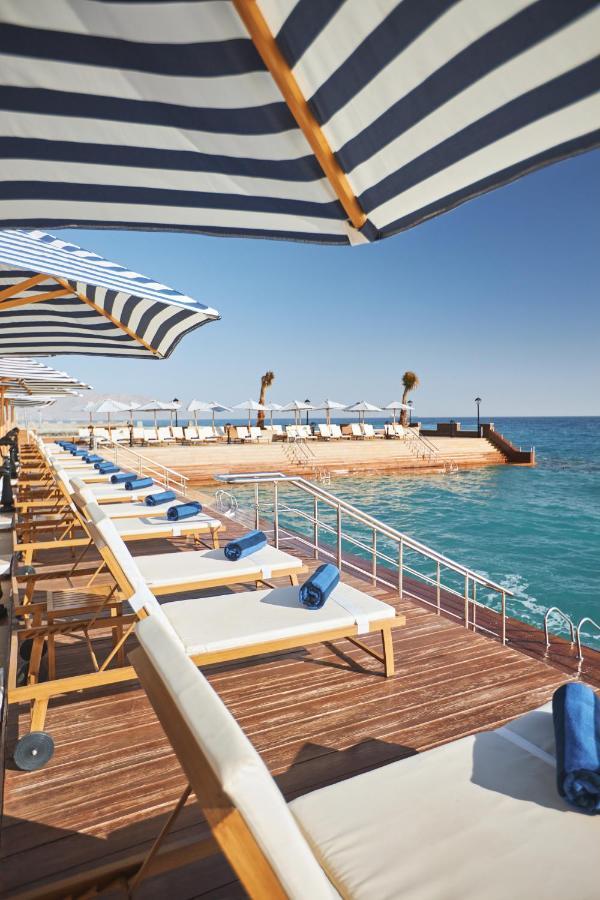 Tolip Resort El Galala Majestic Ajn-Szuhna Kültér fotó The photo depicts a serene outdoor area by the waterfront. There are several lounge chairs arranged in a row, each equipped with blue towels. Large, striped umbrellas provide shade over some of the chairs. In the background, there is a wooden deck th