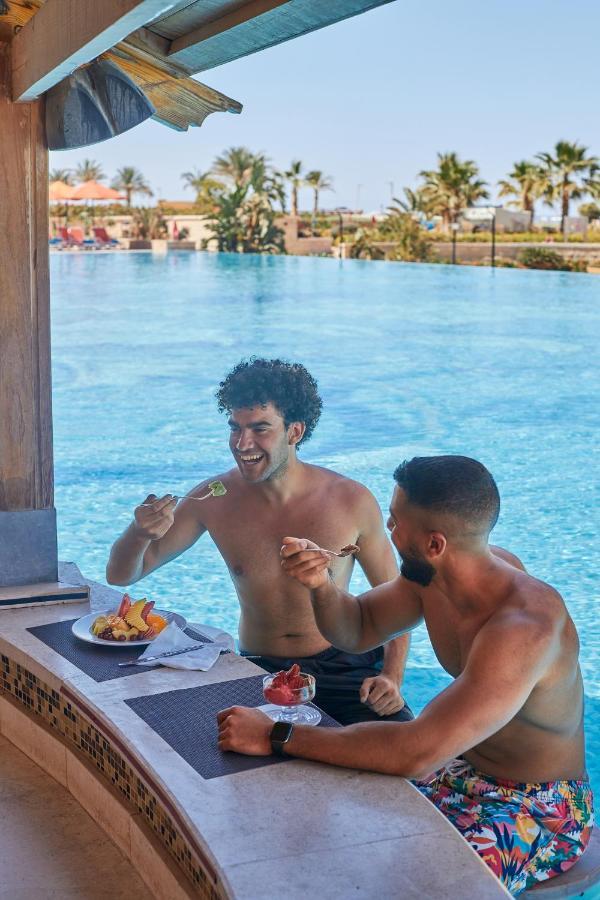 Tolip Resort El Galala Majestic Ajn-Szuhna Kültér fotó The photo features two young men enjoying a meal together. They are sitting at a poolside table with a clear blue swimming pool in the background. The men are shirtless, suggesting a casual, relaxed atmosphere, likely in a warm environment. One man i