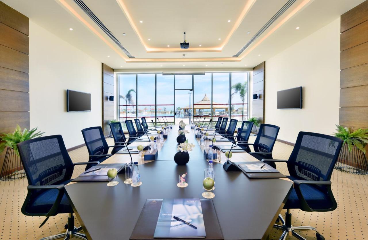 Tolip Resort El Galala Majestic Ajn-Szuhna Kültér fotó The image shows a modern conference room with a large, dark rectangular table surrounded by ergonomic black chairs. The room features large windows that allow natural light to enter and provide a view of the outdoors, including some greenery. On the 