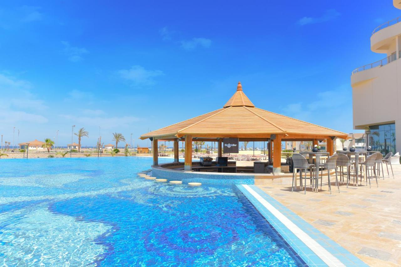 Tolip Resort El Galala Majestic Ajn-Szuhna Kültér fotó The photo shows a beautiful outdoor setting featuring a large swimming pool with clear blue water. In the center, there is a thatched-roof gazebo, which serves as a shaded seating area. Surrounding the gazebo are lounge chairs and a bar area with hig