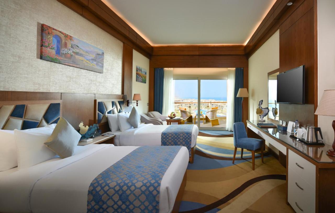 Tolip Resort El Galala Majestic Ajn-Szuhna Kültér fotó The photo shows a well-decorated hotel room featuring two beds with decorative pillows. The room has wooden accents and modern furnishings, including a desk, a television, and lamps. Large windows flood the space with natural light, offering a view o