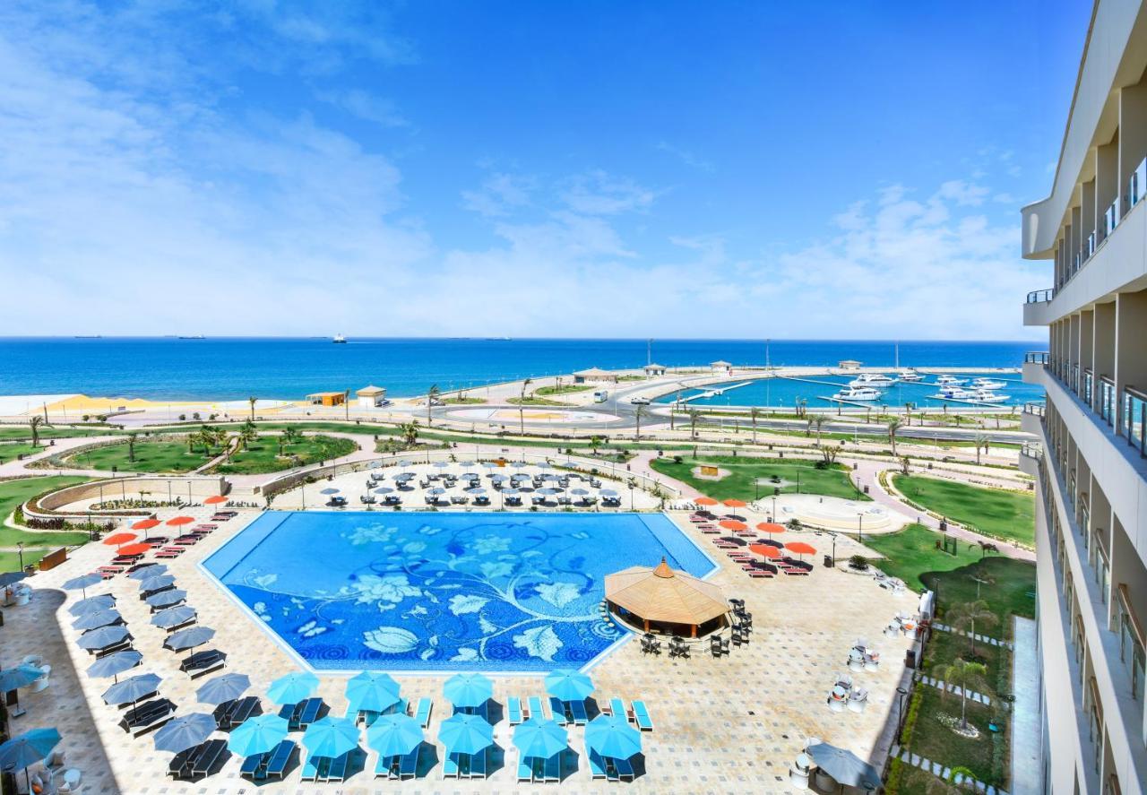 Tolip Resort El Galala Majestic Ajn-Szuhna Kültér fotó The photo shows a beautiful outdoor view of a resort area featuring a large swimming pool with a decorative underwater design. Surrounding the pool are numerous sun loungers and umbrellas in a variety of colors. In the background, there is a scenic c