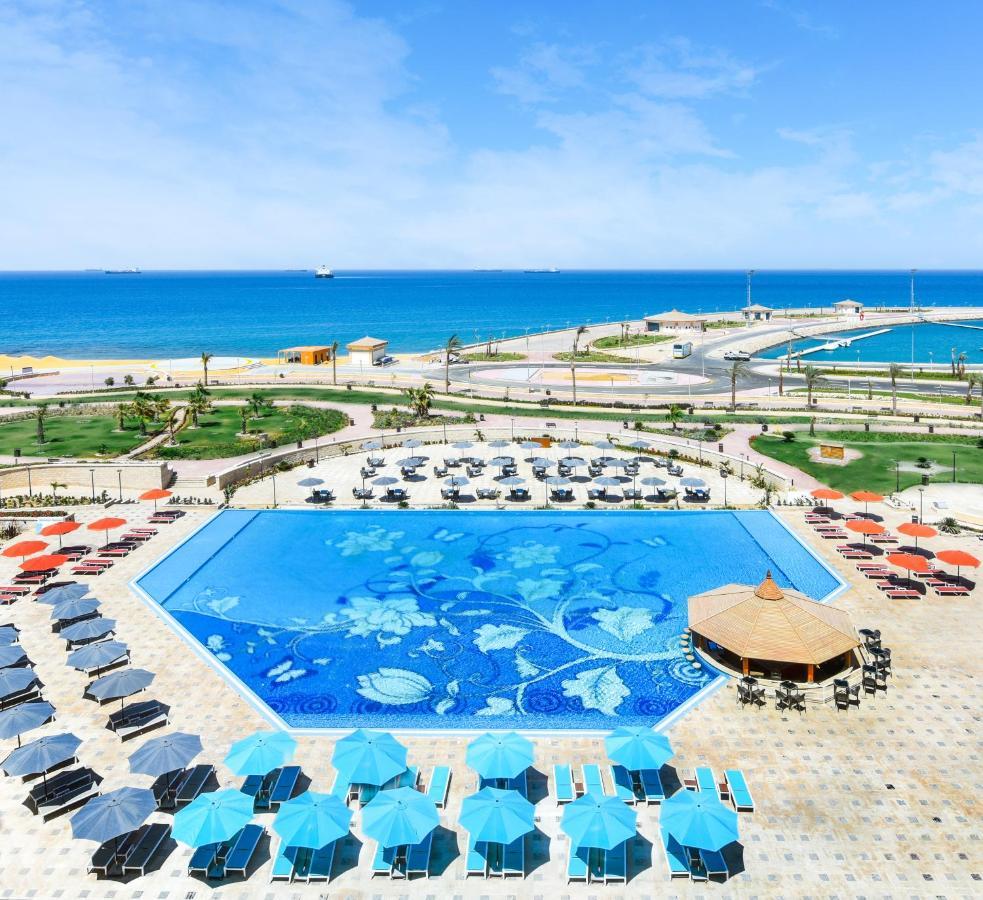 Tolip Resort El Galala Majestic Ajn-Szuhna Kültér fotó The photo features a large swimming pool with a decorative design on its surface, surrounded by sun loungers and umbrellas. In the background, there is a view of the sea, with a clear blue sky and a few scattered clouds. The area is well-maintained, 