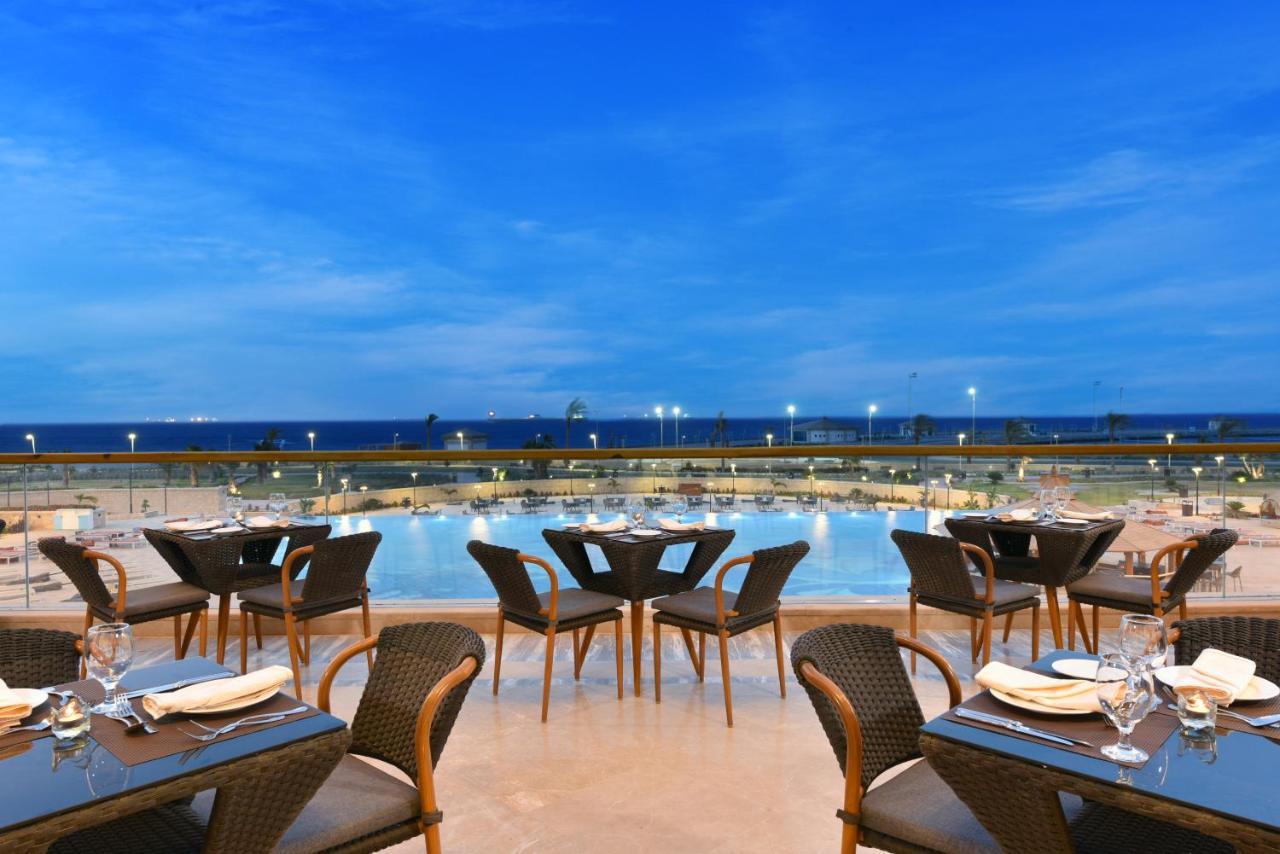 Tolip Resort El Galala Majestic Ajn-Szuhna Kültér fotó The photo shows an outdoor dining area with several neatly arranged tables and chairs. The tables are elegantly set with cutlery and plates, suggesting a fine dining experience. In the background, there is a large swimming pool that reflects the blue