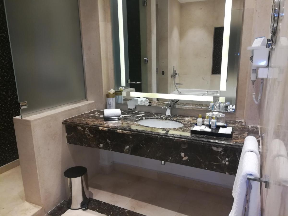 Tolip Resort El Galala Majestic Ajn-Szuhna Kültér fotó The photo shows a modern bathroom with elegant design features. There is a large vanity made of dark marble with a sink and a set of toiletries neatly arranged on top. A mirror with built-in lighting is mounted above the sink. To the side, there is a
