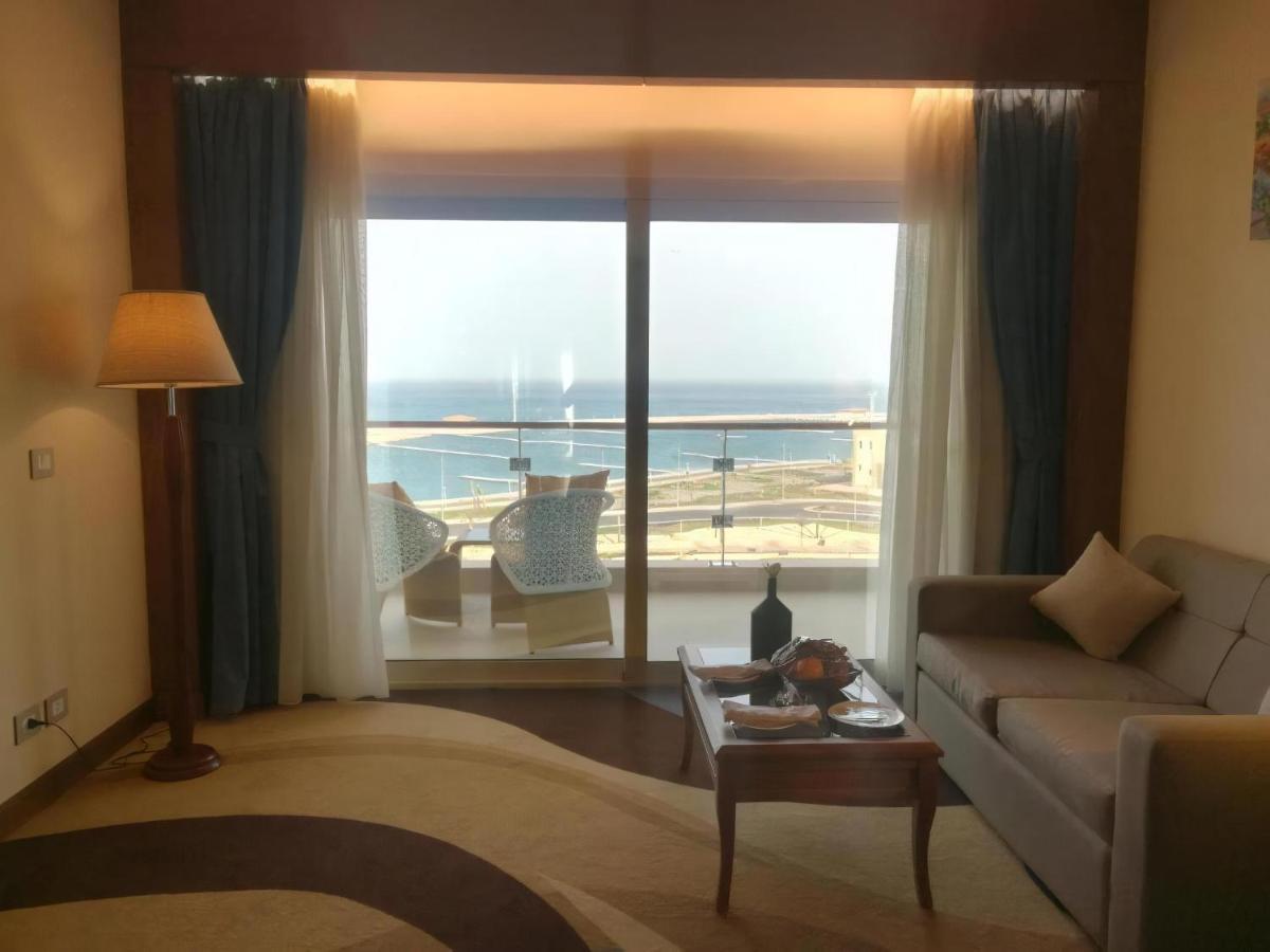 Tolip Resort El Galala Majestic Ajn-Szuhna Kültér fotó The photo shows a stylish interior of a room with a large window overlooking the sea. The window features sheer and thick curtains and is framed by a balcony with two chairs visible outside. Inside, there is a comfortable-looking sofa, a coffee table