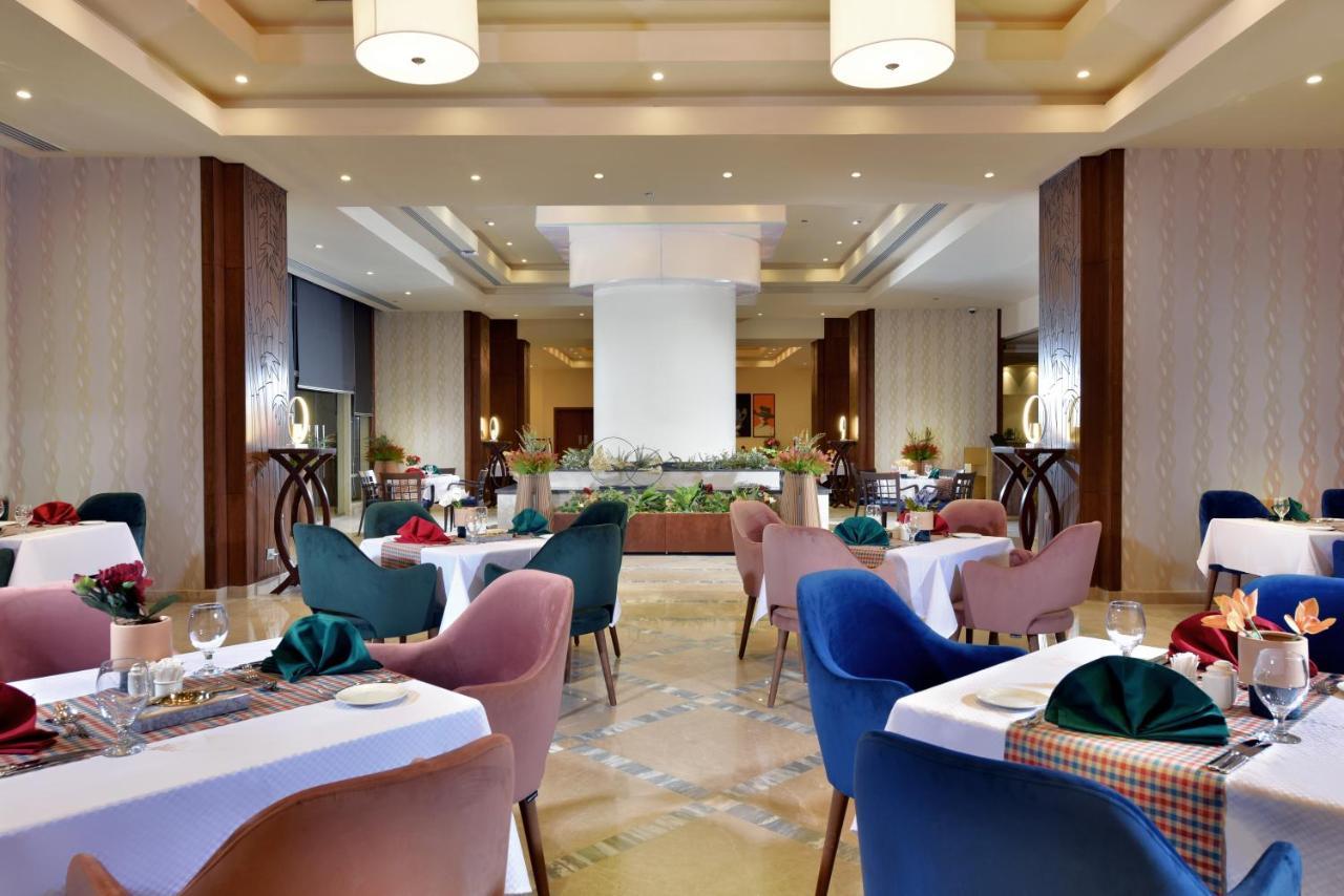 Tolip Resort El Galala Majestic Ajn-Szuhna Kültér fotó The photo shows a spacious and elegantly designed restaurant interior. The layout includes several round tables dressed in white tablecloths, each set with decorative plates, cutlery, and colorful napkins. The chairs are upholstered in a variety of c