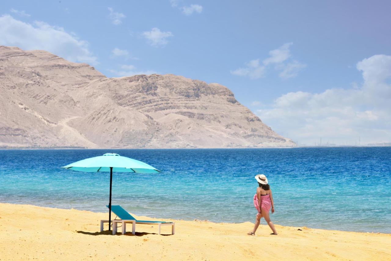 Tolip Resort El Galala Majestic Ajn-Szuhna Kültér fotó The photo depicts a serene beach scene. In the foreground, there is a sandy shore with a single beach chair and an umbrella in a light blue color. A person dressed in a pink outfit is walking along the shoreline, enjoying the beach atmosphere. In the